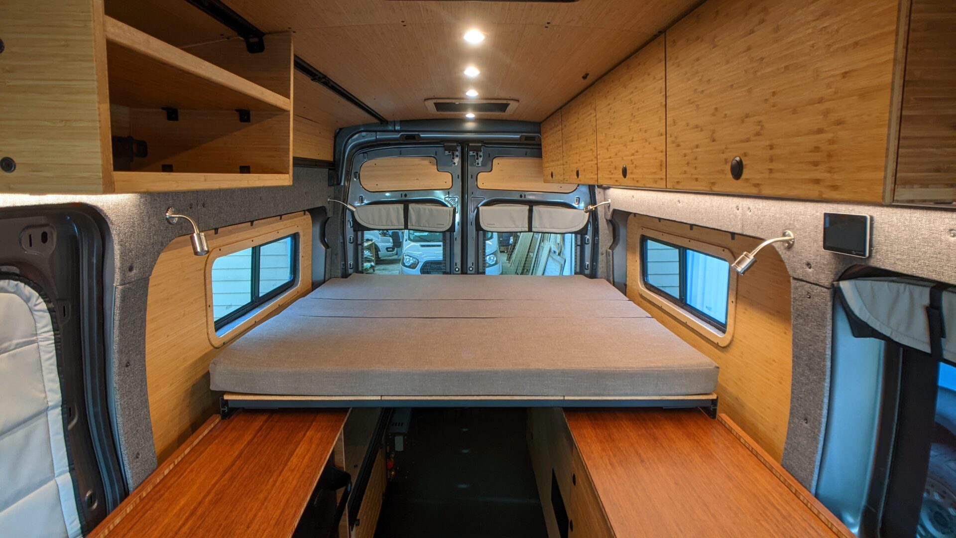 Interior of a camper van with oak plywood overhead cabinets, a small desk, and built‑in lighting along the wall edge.