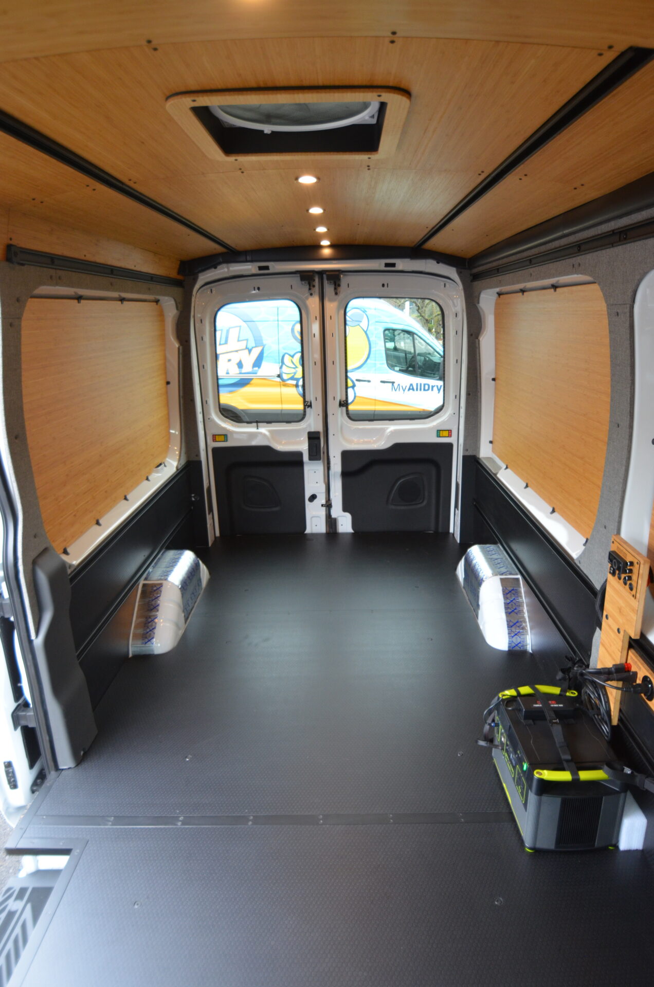 Interior of a cargo van with wooden panel walls and ceiling, back double doors, and a black rubberized floor; LED lights along the ceiling and a ceiling vent visible at top center.